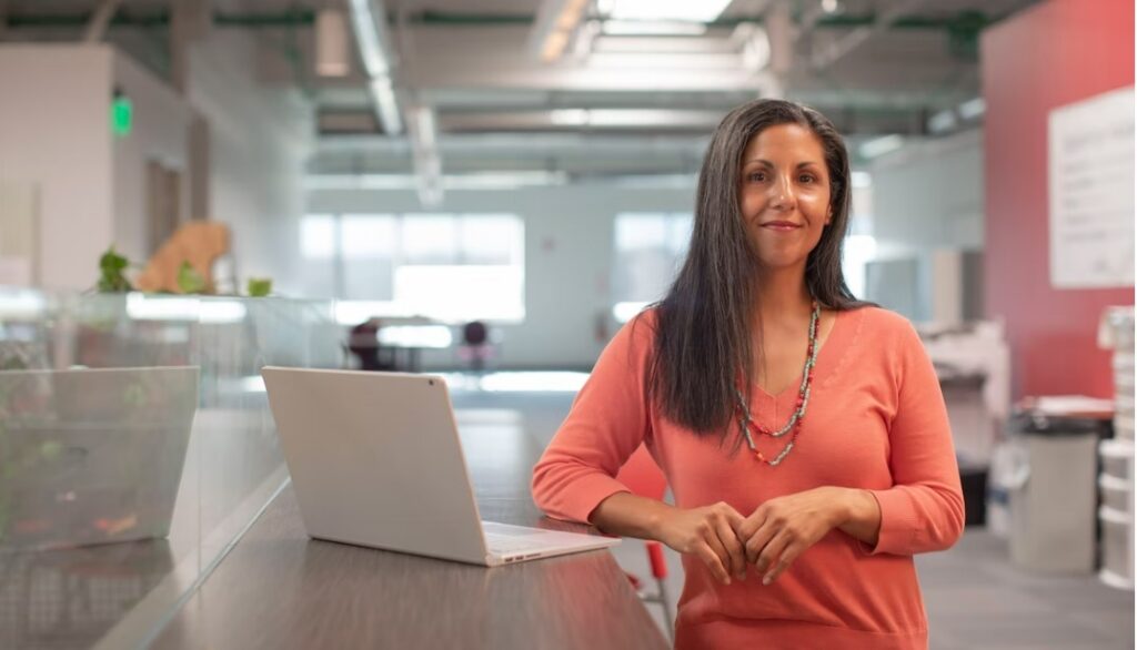 A woman of colour wearing a salmon coloured jumper, standing with a laptop in a large open plan office 