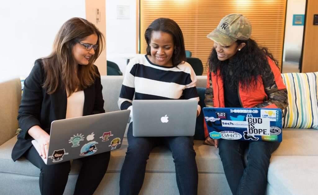 Three women of colour sitting on sofas and looking at each other's laptops 