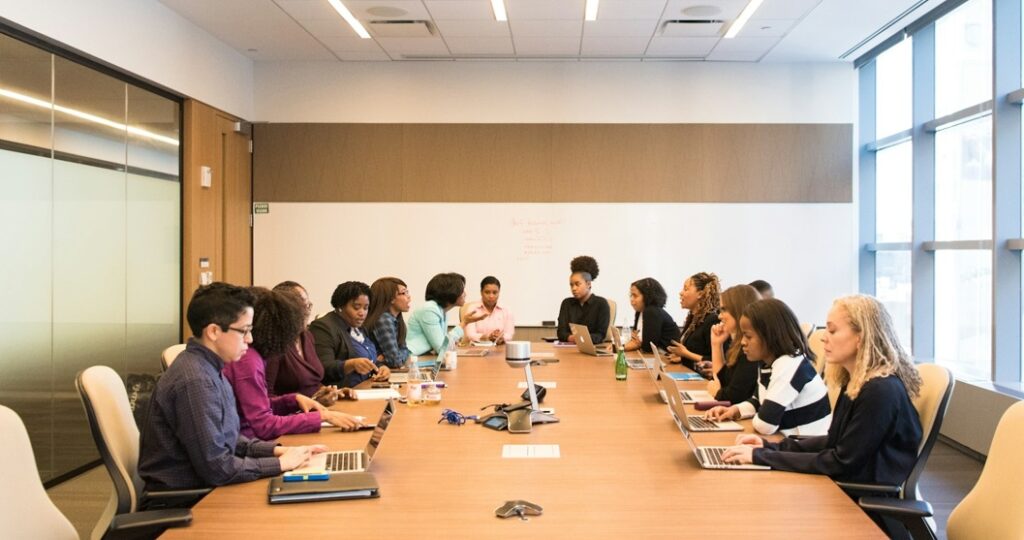 A group of people with representation from marginalised communities sitting around a large wooden table in a modern boardroom having a meeting 